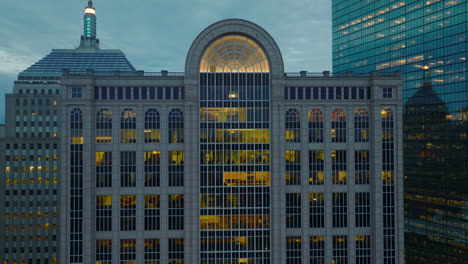 Pull-back-shot-of-illuminated-interior-of-high-rise-commercial-building-at-dusk.-Backwards-reveal-of-surrounding-downtown-office-buildings.-Boston,-USA