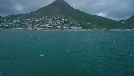 Fly-above-water-surface-along-sea-coast.-Watching-whales-in-wildlife.-Residential-buildings-and-mountain-in-background.
