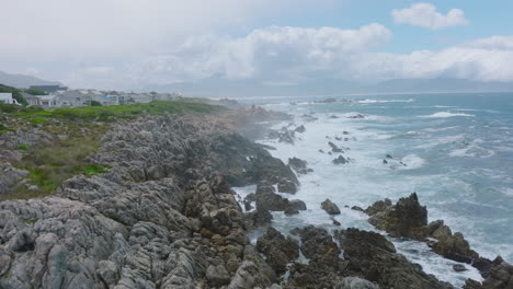 Fly-above-waves-crashing-to-coastal-rocks.-Backwards-reveal-of-rugged-and-jagged-rocky-sea-coast-near-residential-development.