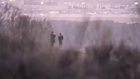 back-view-of-couple-taking-photos-with-the-smartphone-on-top-of-a-hill-with-airport-and-city-as-background-foreground-bushes